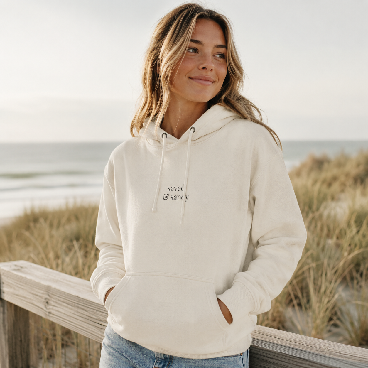 Woman wearing a cream Saved and Sandy embroidered hoodie near the beach